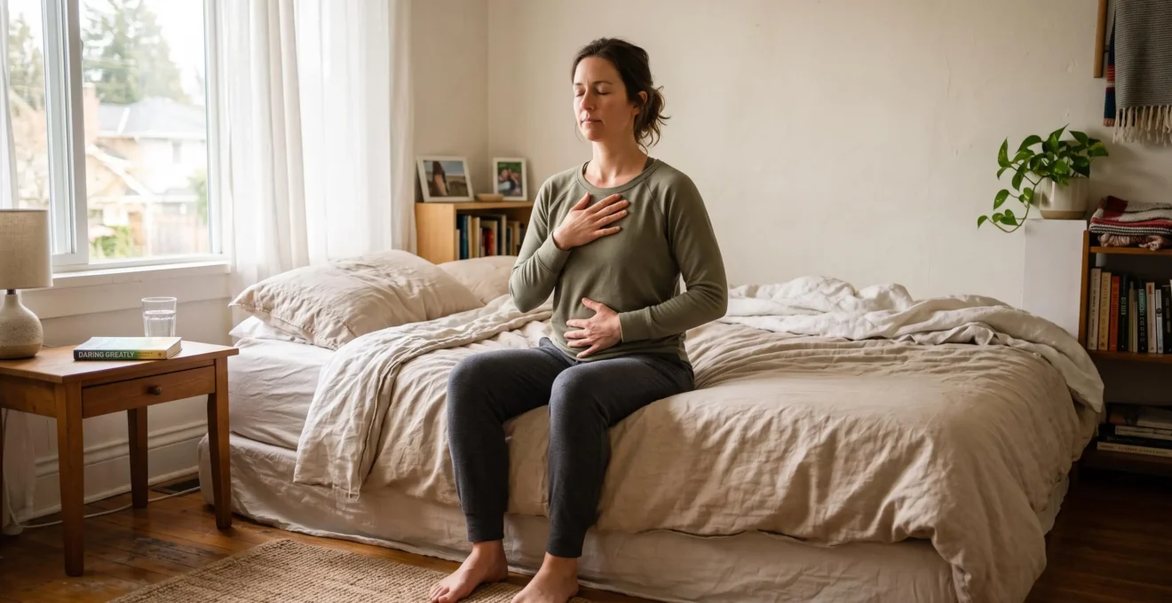 Une femme assise au bord de son lit pratique la respiration ventrale, une main posée sur le ventre et l'autre sur la poitrine, dans une chambre baignée de lumière matinale.
